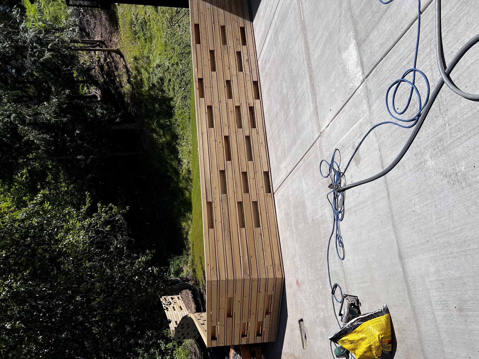 Long custom horizontal-slat wood fence under construction running alongside a residential concrete driveway, with sun and shade across the slats